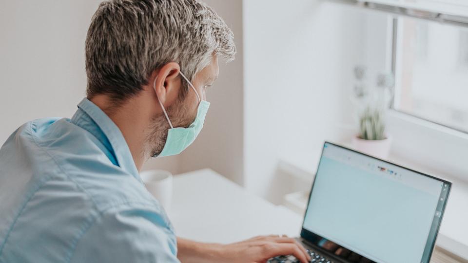 Ein Mann sitzt mit einer medizinischen Mund- und Nasenschutzmaske schreibend vor einem aufgeklappten Laptop.
