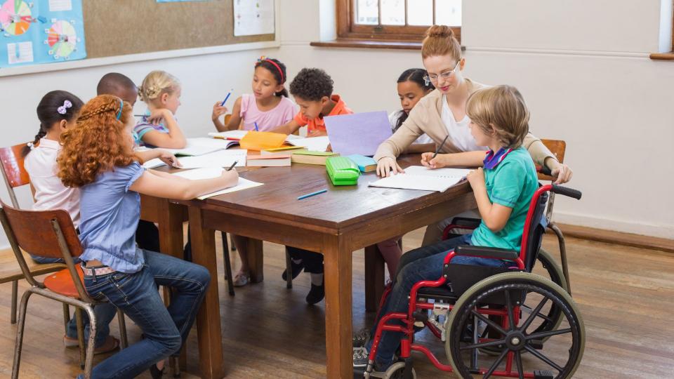 Ein Klassenraum mit mehreren Schülern an einem Tisch. Eine Lehrerein hilft einem Kind im Rollstuhl