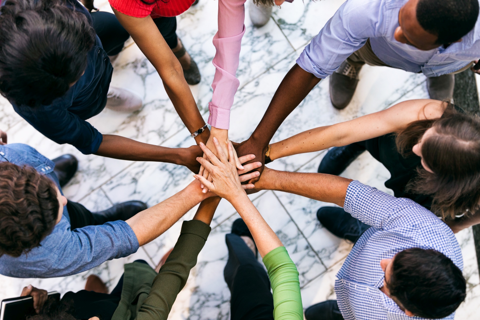 Eine Gruppe von jungen Erwachsenen, die unterschiedlicher ethnischer Herkunft sind und gemeinsam in einem geschlossenen Kreis stehen, in dessen Mitte sich die Hände der Personen berühren.