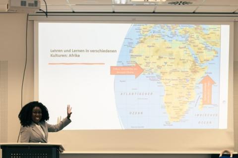 Abgebildet ist eine Frau, die etwas zu dem afrikanischen Kontinent erklärt, der an einer Leinwand zu erkennen ist.