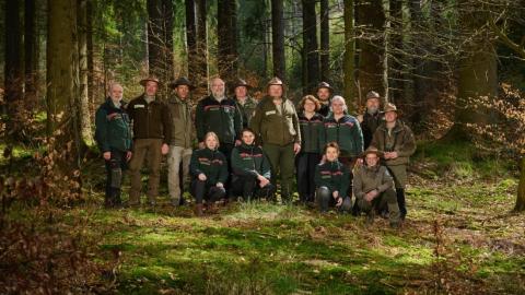 Abgebildet sind Waldpädagoginnen und Waldpädagogen, Rangerinnen und Ranger, Försterinnen und Förster sowie Verwaltungsexpertinnen und Verwaltungsexperten, die sich in einem Waldgebiet befinden. 