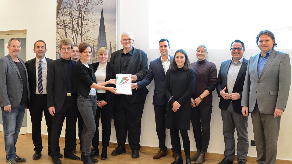 Regierungspräsident Hans-Josef Vogel (Mitte) überreicht Projektleiterin Alessa Näpel (5.v.l.) und den anderen Projektbeteiligten den Förderbescheid.