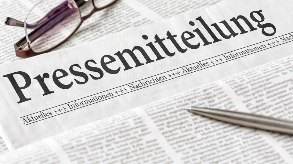 Abgebildet sind mehrere übereinanderliegende Zeitungen. Im Fokus steht dabei ein Zeitungsartikel mit dem Titel Pressemitteilung. Darüber hinaus liegen ein Kugelschreiber und eine Lesebrille auf den Zeitungen. 