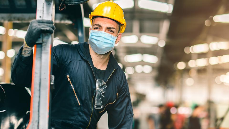 Abgebildet ist ein Mann in Arbeits- beziehungsweise Schutzkleidung, der sich in einer großen Fabrikhalle befindet. Der Mann trägt darüber hinaus eine Mund- und Nasenschutzmaske.