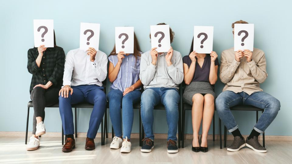 Drei Frauen und drei Männer sitzen nebeneinander auf Stühlen vor einer hellblauen Wand. Vor ihren Gesichtern halten sie jeweils ein weißes Blatt Papier, auf dem ein dunkles Fragezeichen abgebildet ist.
