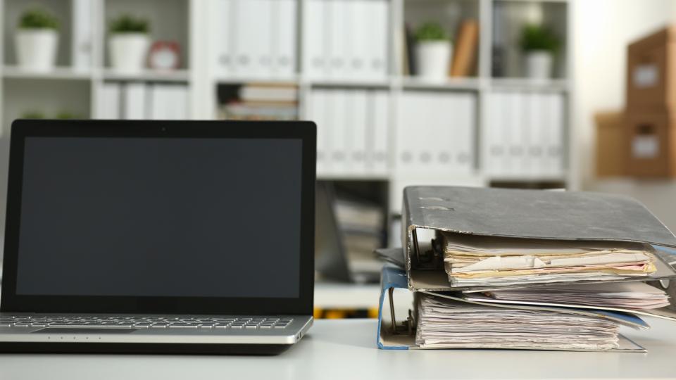 Ein aufgeklappter Laptop und zwei übereinandergestapelte Aktenordner auf einen Schreibtisch.