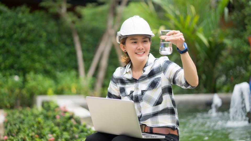 Eine Frau sitzt in einer Grünanlage und schaut auf ein mit Wasser gefülltes Reagenzglas. Sie trägt einen Schutzhelm und auf ihrem Schoß befindet sich ein Laptop.
