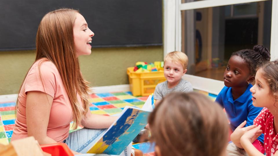 Vier Kinder mit unterschiedlicher ethnischer Herkunft hören aufmerksam einer Erzieherin zu, die aus einem Kinderbuch vorliest.