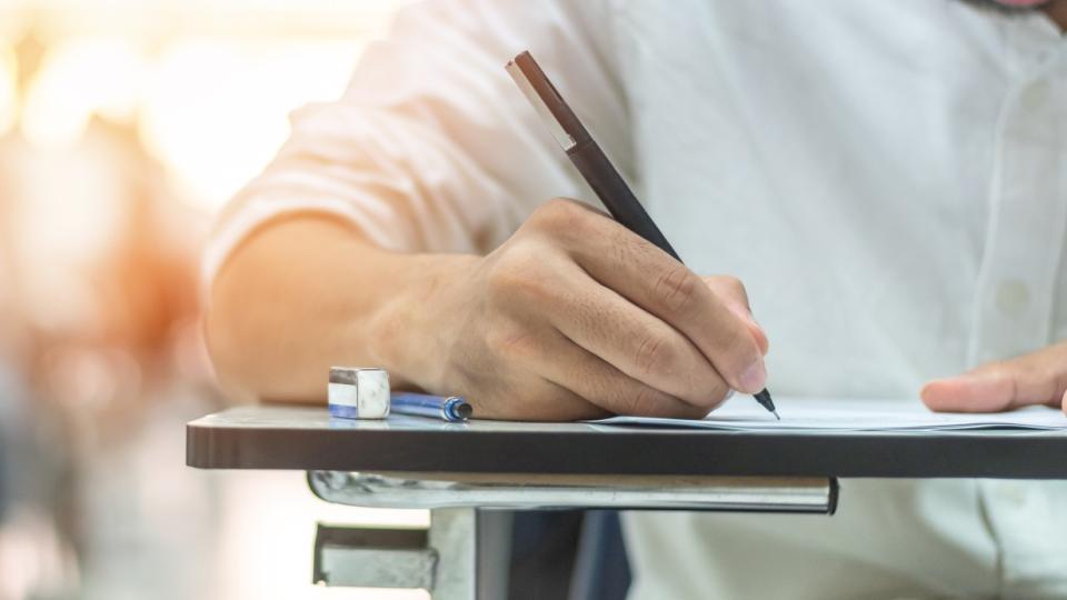 Im Vordergrund sitzt eine Person in einem Klassenraum und schreibt auf ein Blatt Papier. Die Schülerinnen und Schüler im Hintergrund sitzen an Einzeltischen.