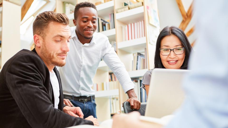 Eine Gruppe von jungen Erwachsenen, die unterschiedlicher ethnischer Herkunft sind und gemeinsam in einem Büro oder einer Bibliothek lernen. Auf dem Tisch befindet sich ein Buch und ein aufgeklappter Laptop.