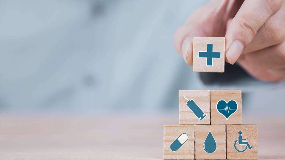 Eine Person stapelt kleine Holzquadrate zu einer Pyramide. Auf den Quadraten sind medizinische Symbole abgebildet, wie z. B. ein Medikament oder eine Spritze, aber auch eine Person im Rollstuhl.