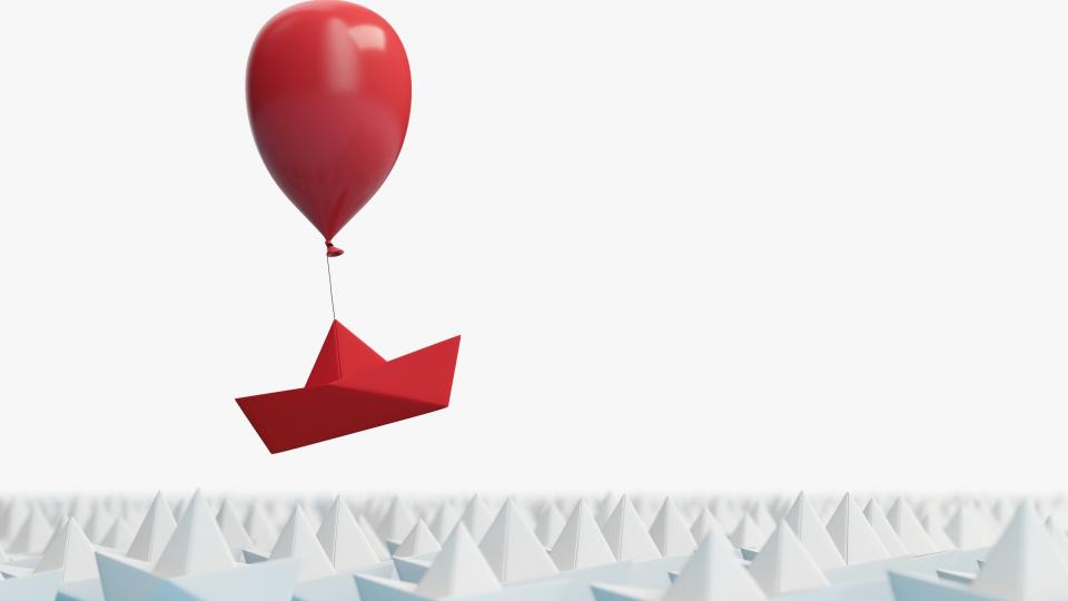 Ein Ausschnitt von hunderten weißen Papierbooten, die in perfekter Reihung neben- beziehungsweise voreinander stehen. Darüber befindet sich ein rotes Papierboot, das an einem roten Luftballon hängend davon schwebt.