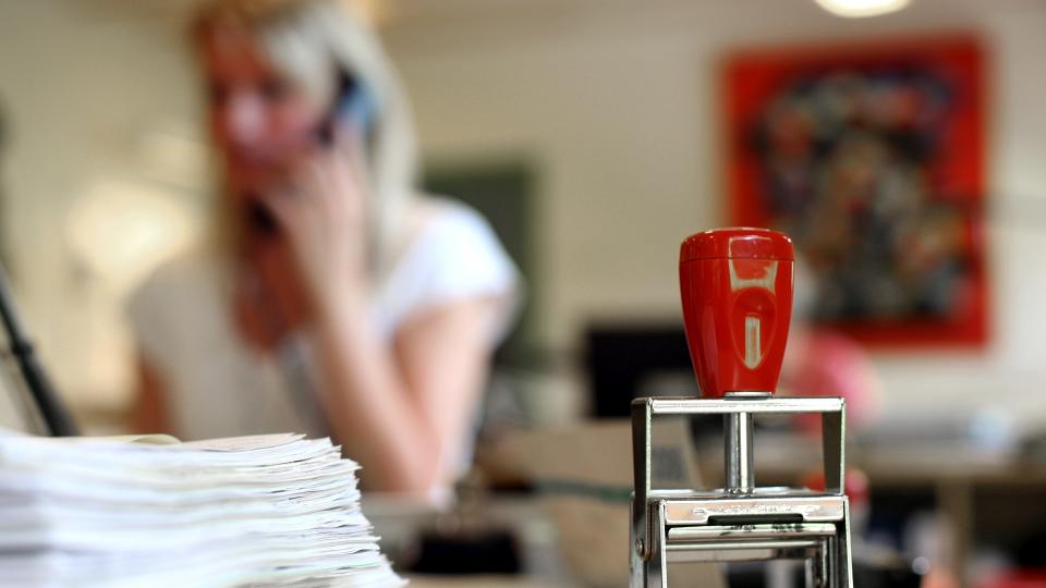Im Fokus steht ein Stempel neben einem Blätterstapel und im Hintergrund sitzt eine telefonierende Frau an ihren Arbeitsplatz.