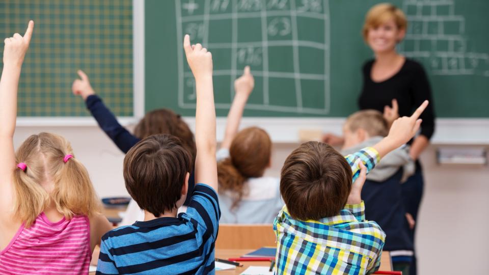 Mehrere Grundschulkinder befinden sich in einem Klassenraum und machen ein Handzeichen, welches andeutet, dass sie der an der Tafel stehenden Lehrerin etwas sagen wollen. 