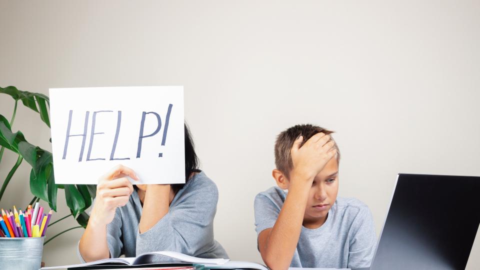 Eine Mutter sitzt mit ihrem Kind an einem Schreibtisch, auf dem sich Schulunterlagen und ein Laptop befinden. Der Junge sieht verzweifelt aus und stützt seinen Kopf mit seiner Hand. Die Mutter hält ein Blatt Papier mit der Aufschrift Help! (zu Deutsch: Hilfe!) hoch.