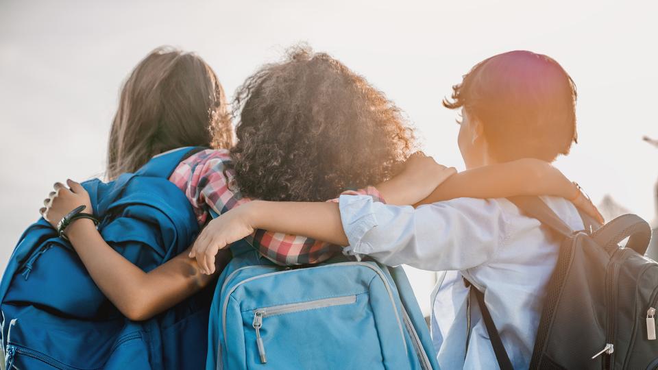Drei eng beieinander stehende Kinder, die Schulranzen tragen und in die Ferne schauen.
