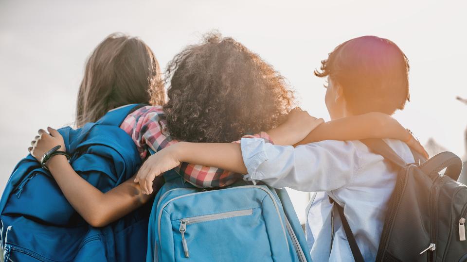 Drei eng beieinander stehende Kinder, die Schulranzen tragen und in die Ferne schauen. 