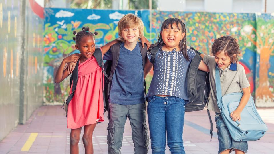 Vier lachende und freundschaftlich nebeneinander stehende Kinder, die unterschiedlicher ethnischer Herkunft sind. Im Hintergrund steht eine farbenfroh bemalte Wand. 