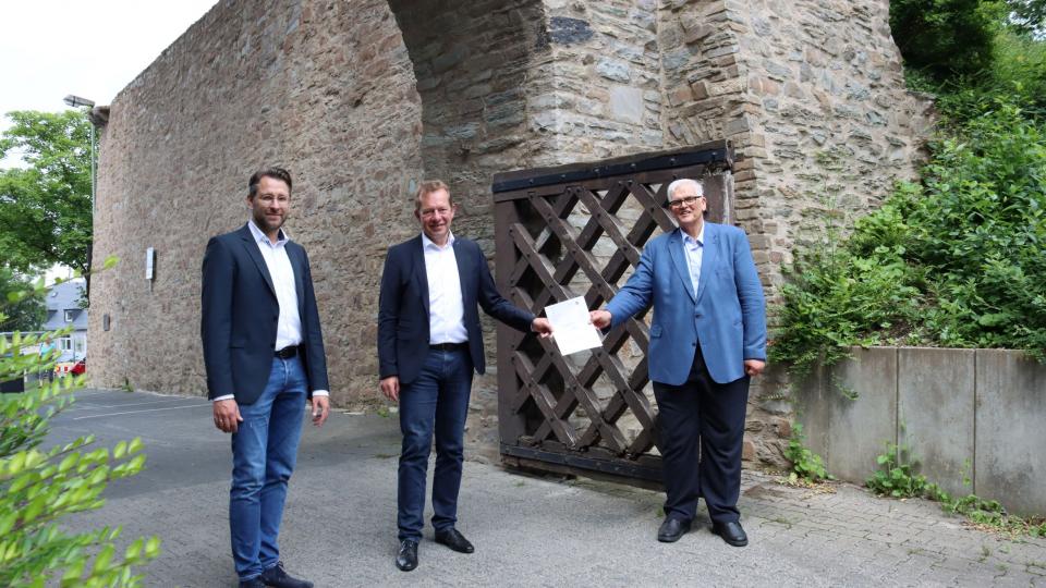 Der Siegener Stadtbaurat Henrik Schumann (l.), Bürgermeister Steffen Mues (m.) und Regierungspräsident Hans-Josef Vogel (r.) bei der Bescheid-Übergabe vor der bereits sanierten Stadt- und Schlossmauer im Bereich „Hasengarten“ 