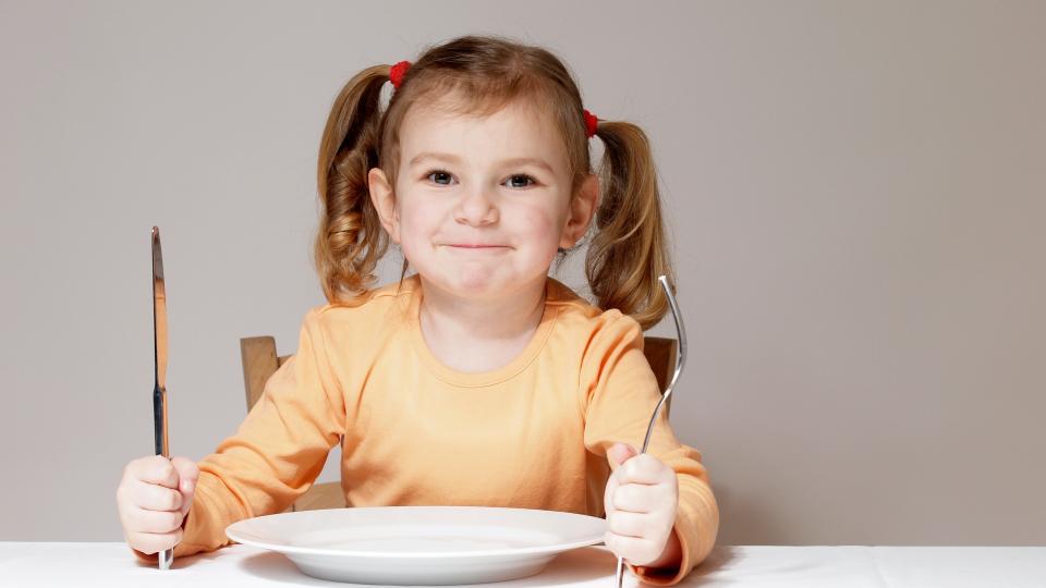 Ein kleines Mädchen sitzt mit Messer und Gabel in den Händen vor einem leeren Teller.