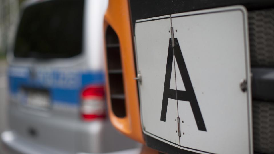 Das Bild zeigt ein Schild "A" an der Front eines LKW, im Hintergrund ist verschwommen ein Polizeiwagen zu sehen.