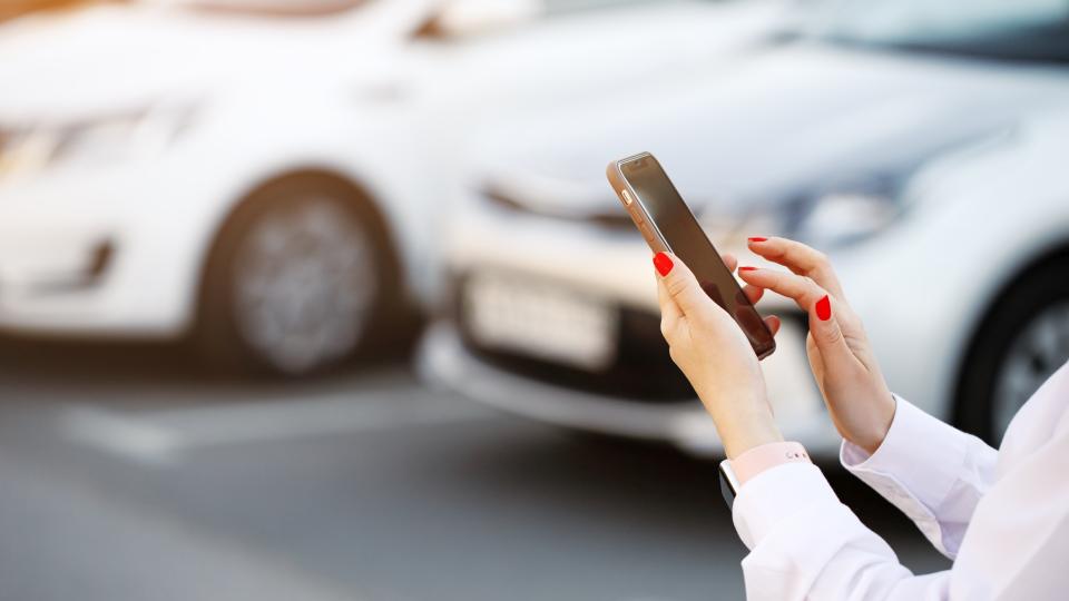 Nahaufnahme von Händen einer Frau mit Smartphone vor parkenden Autos.