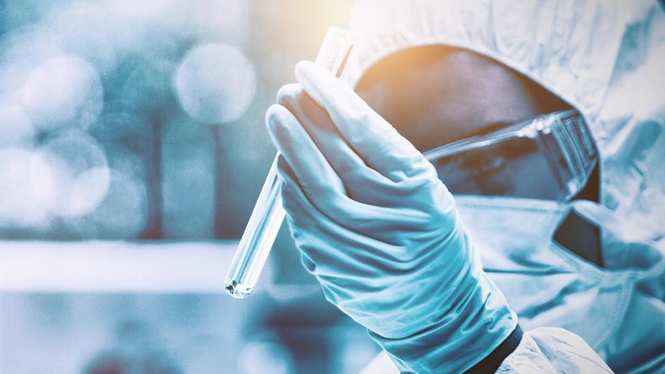 Eine Person in Schutzkleidung hält ein Reagenzglas auf Augenhöhe in einer Hand. Die Schutz- beziehungsweise Laborkleidung besteht aus einem chemikalienbeständigen Anzug, einer Schutzbrille und Gummihandschuhen.