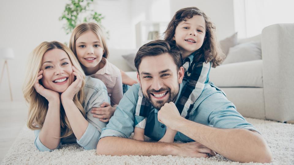Abgebildet ist eine glückliche Familie. Die Eltern und die beiden Kinder liegen auf dem Teppich ihres Wohnzimmers.