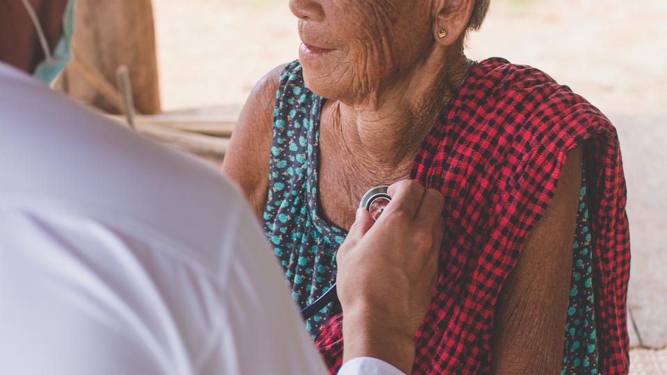 Ein Arzt untersucht mit Hilfe eines Stethoskops eine Seniorin mit Migrationshintergrund. 