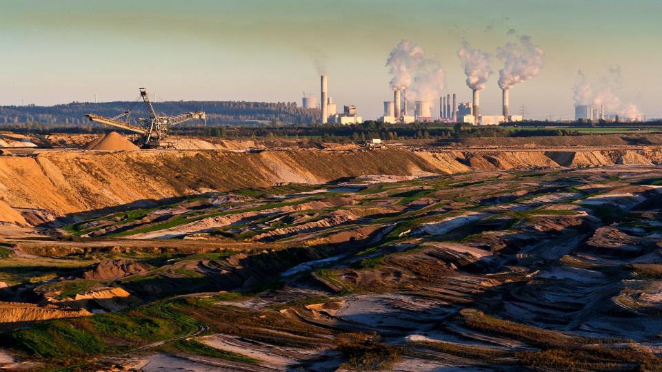 Im Vordergrund ist ein Tagebau abgebildet und im Hintergrund ein Industriegebiet mit mehreren Schornsteinen, die sehr viel hellen Rauch an die Umgebung abgeben. 