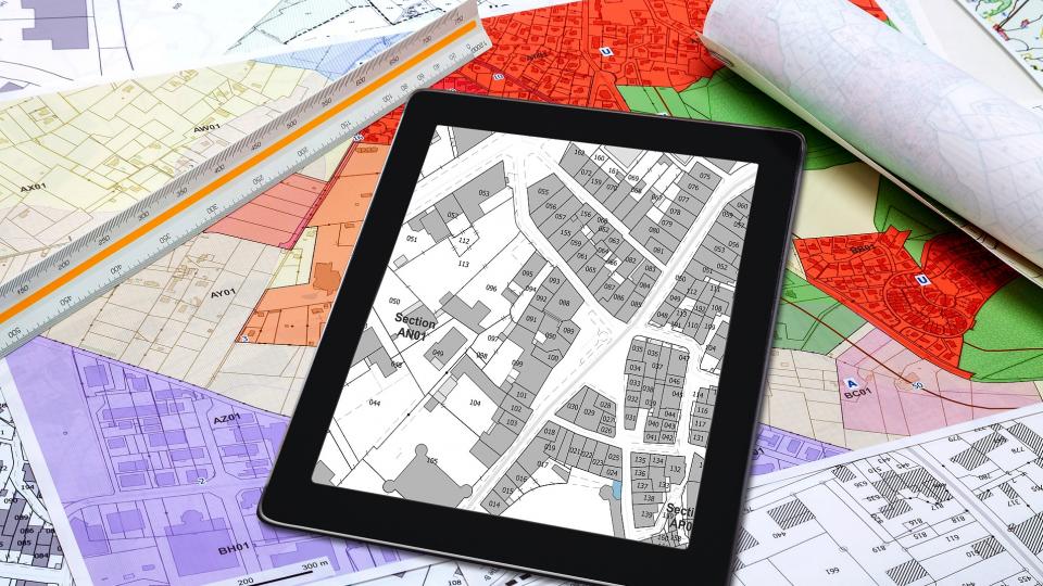 Abgebildet sind mehrere Lagepläne in Papierform und ein Lageplan auf einem Tablet sowie ein Dreikantlineal.
