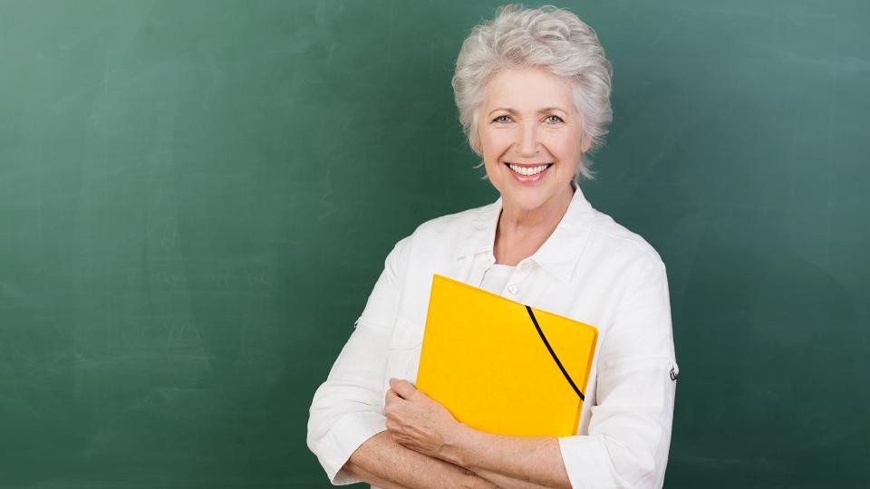Eine ältere Dame steht lächelnd vor einer Tafel. In den Händen hält sie einen gelben Ordner. 