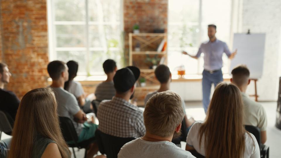 Ein Redner steht mit ausgebreiteten Armen vor jungen Erwachsenen. Hinter dem Redner steht ein Flipchart. 