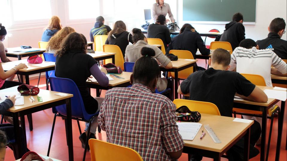Schülerinnen und Schüler sitzen an Einzeltischen in einem Klassenraum und schreiben eine Prüfung. Vor den Schülerinnen und Schülern steht eine Aufsichtsperson. 
