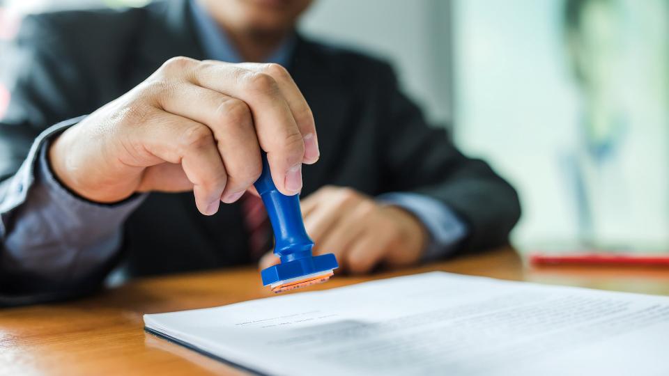 Ein Mann hält einen blauen Stempel in der Hand und deutet an, ein Schriftstück stempeln zu wollen. 