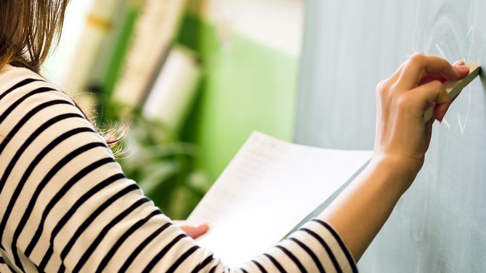 Eine Lehrerin steht vor einer Tafel. In der linken Hand hält sie ein Blatt Papier und mit der rechten Hand schreibt sie mit Kreide an die Tafel. 