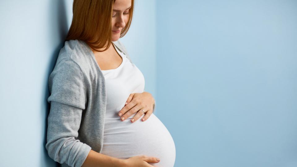 Eine blonde, schwangere Frau lehnt an einer blauen Wand