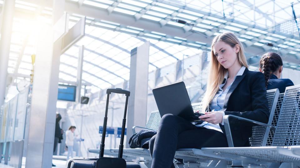 Eine blonde Frau sitzt mit Ihrem Laptop in der Hand auf einer Bank in einem Bahnhofsgebäude 