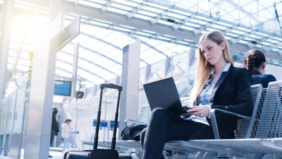 Eine blonde Frau sitzt mit Ihrem Laptop in der Hand auf einer Bank in einem Bahnhofsgebäude