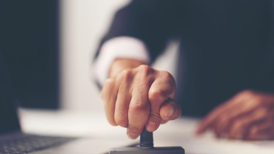 Eine Nahaufnahme von einer Hand die einen breiten Stempel auf ein Dokument drückt. Im Hintergrund ist ein Laptop zu sehen