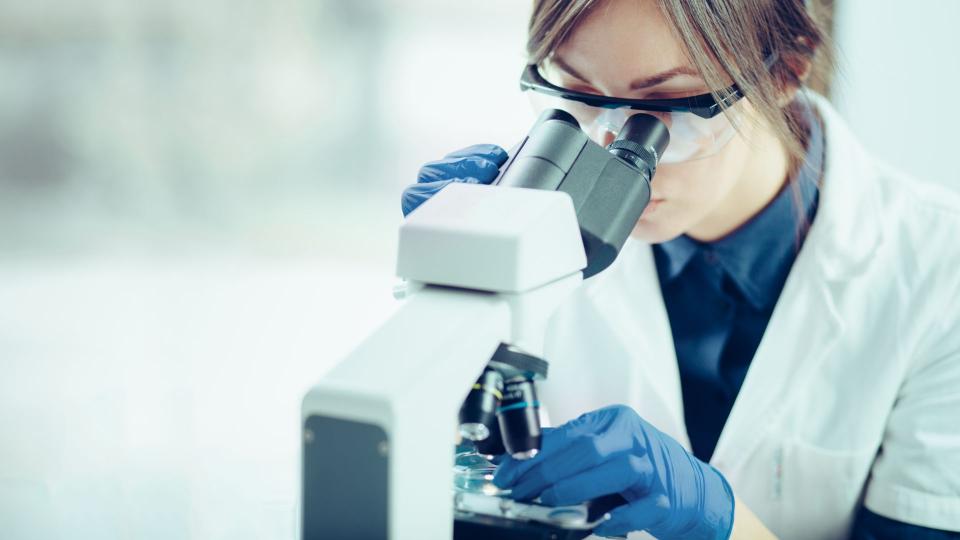 Eine Frau in einem weißen Kittel schaut durch eine Schutzbrille hindurch in eine großes Mikroskop