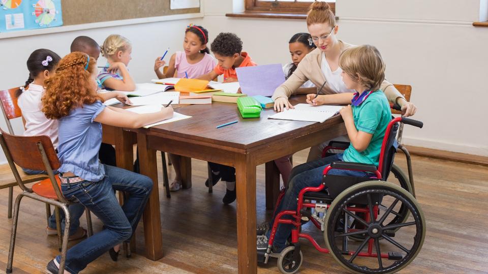 Grundschülerinnen und Grundschüler sitzen geschlossen an zwei aneinander geschobenen Tischen. Eines der Kinder sitzt in einem Rollstuhl und wird von einer Lehrerin bei der Bearbeitung von Schulaufgaben betreut. Die anderen Kinder beschäftigen sich ebenfalls mit Schulaufgaben. 