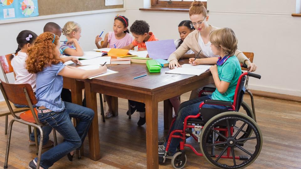 Grundschülerinnen und Grundschüler sitzen geschlossen an zwei aneinander geschobenen Tischen. Eines der Kinder sitzt in einem Rollstuhl und wird von einer Lehrerin bei der Bearbeitung von Schulaufgaben betreut. Die anderen Kinder beschäftigen sich ebenfalls mit Schulaufgaben.