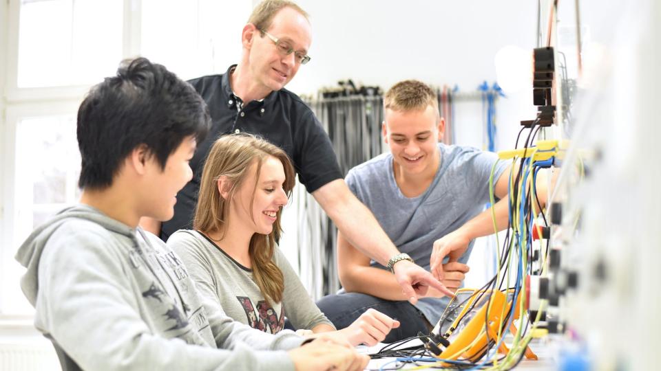 3 Schüler lassen sich von einem Lehrer in einem Technikraum einen Stromzähler erklären