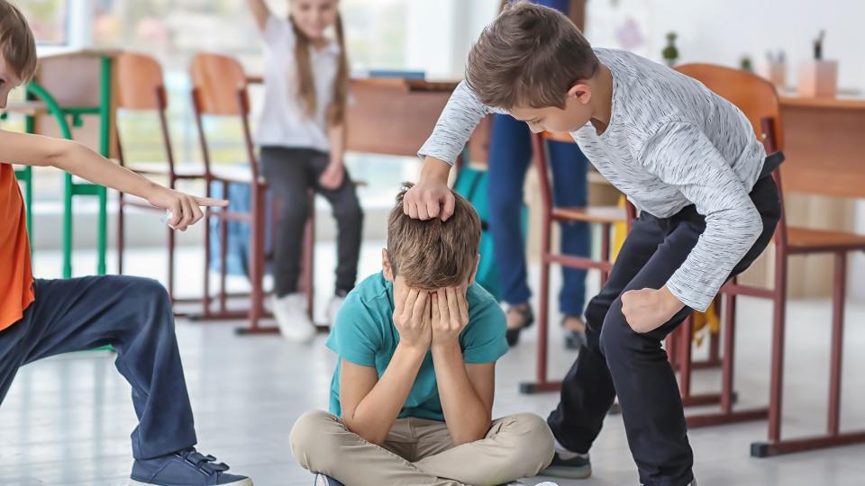 Ein auf dem Boden sitzender und weinender Junge wird von seinen Klassenkameraden gemobbt. Ein Junge zeigt mit dem Finger auf ihn und ein anderer deutet einen Faustschlag an. Ein Mädchen im Hintergrund befürwortet die Aktion ihrer Klassenkameraden. 