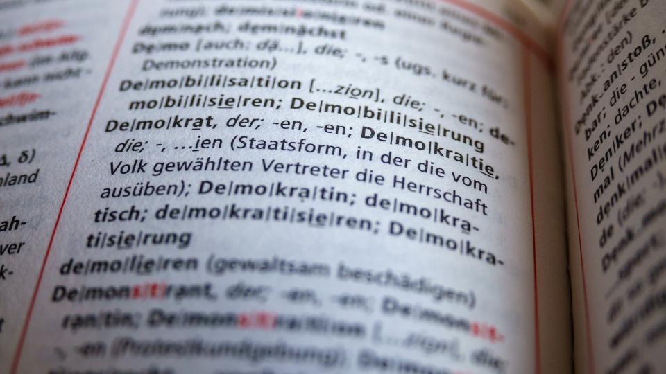 Abgebildet ist ein Ausschnitt aus dem Duden, in dem das Wort "Demokratie" im Vordergrund steht. 