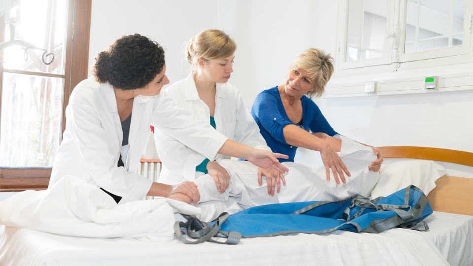 Ein menschlicher Dummy liegt in einem Krankenbett und wird von drei Frauen in eine seitliche Position gedreht. Zwei Frauen tragen weiße Arztkittel und die Frau ganz rechts in der Abbildung trägt normale Alltagskleidung.
