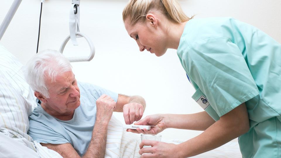 Ein älterer Mann liegt in einem Krankenbett und erhält von einer medizinischen Fachkraft Tabletten und ein Glas Wasser. 