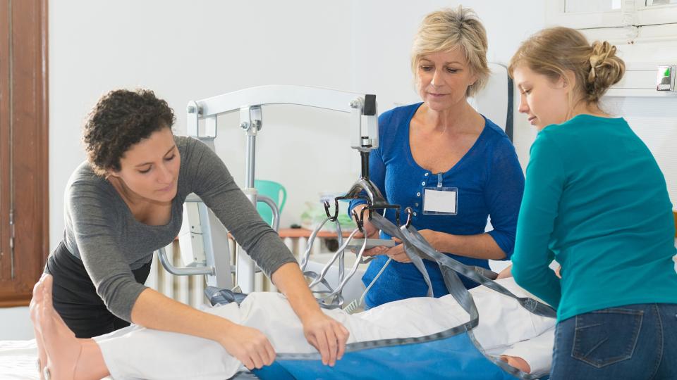 Ein menschlicher Dummy liegt in einem Krankenbett. Drei Frauen befestigen den Dummy an einem Patientenlifter. 