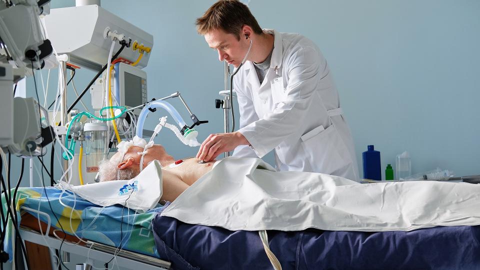Ein Mann liegt in einem Krankenbett und ein medizinisches Beatmungsgerät ist an ihm angeschlossen. Ein Arzt hört dabei den Herzschlag des Mannes mit einem Stethoskop ab.